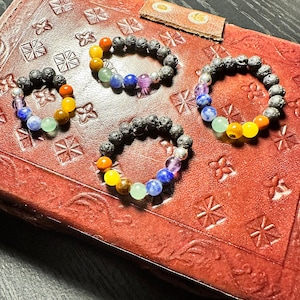 May include: Three chakra bracelets with lava rock beads and colorful gemstone beads. The bracelets are arranged on a brown leather surface.