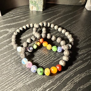 May include: Two black lava rock bracelets with rainbow colored gemstone beads. The bracelets are on a dark wood surface.