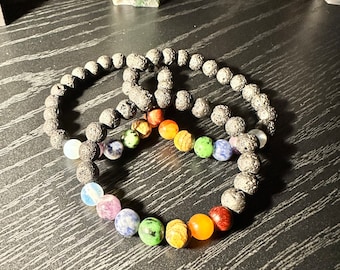 Op de afbeelding: Twee zwarte lava armbanden met regenboogkleurige edelsteen kralen. De armbanden liggen op een donker houten oppervlak.