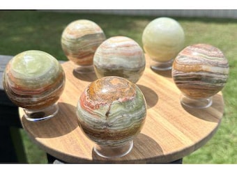 Op de afbeelding: Zes gepolijste stenen bollen met gestreepte patronen in tinten groen, bruin, wit en oranje. Elke bol is glad, rond en rust op een kleine, doorzichtige ronde standaard. De stenen vertonen natuurlijke minerale lagen en gevarieerde kleurwervelingen.