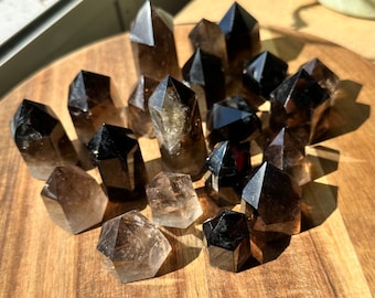 Op de afbeelding: Een verzameling donkere, rookkwarts kristallen punten gerangschikt op een houten oppervlak. De kristallen variëren in grootte en vorm, met scherpe, facetgeslepen punten en een reeks bruine en zwarte tinten. De kristallen worden verlicht door zonlicht.