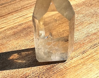 May include: A clear quartz crystal point with a natural, raw finish. The crystal is sitting on a wooden surface.