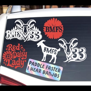 May include: A collection of vinyl decals applied to a car window. The stickers include white goat heads with the text BMFS 33, a red daisy flower with BMFS, a white goat silhouette with BMFS, and a rectangular holographic sticker reading PADDLE FASTER I HEAR BANJOS.