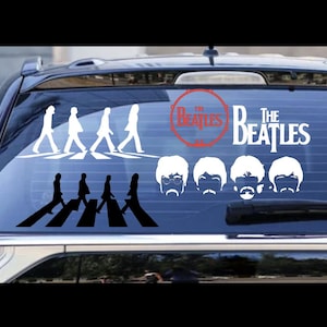 May include: A black car window decal featuring the band The Beatles. The decal includes the band's name, the iconic Abbey Road crossing scene, and the band members' faces. The Beatles logo is in a red circle.