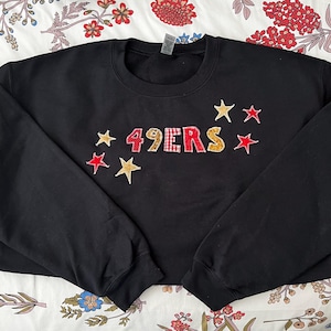 May include: Black cropped sweatshirt with the word "49ERS" in red, gold, and plaid fabric, surrounded by gold and red stars. The sweatshirt has long sleeves and a crew neck.