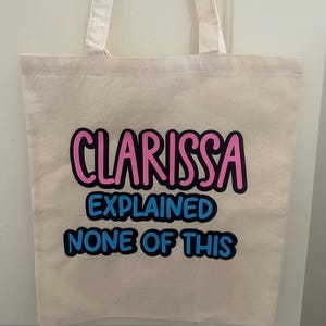 May include: A white canvas tote bag with the text "Clarissa Explained None of This" in pink and blue lettering.