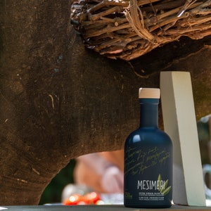 May include: A dark blue glass bottle of Mesimeri Extra Virgin Olive Oil with a white label and handwritten text. The bottle is sitting on a white surface with a wooden background.