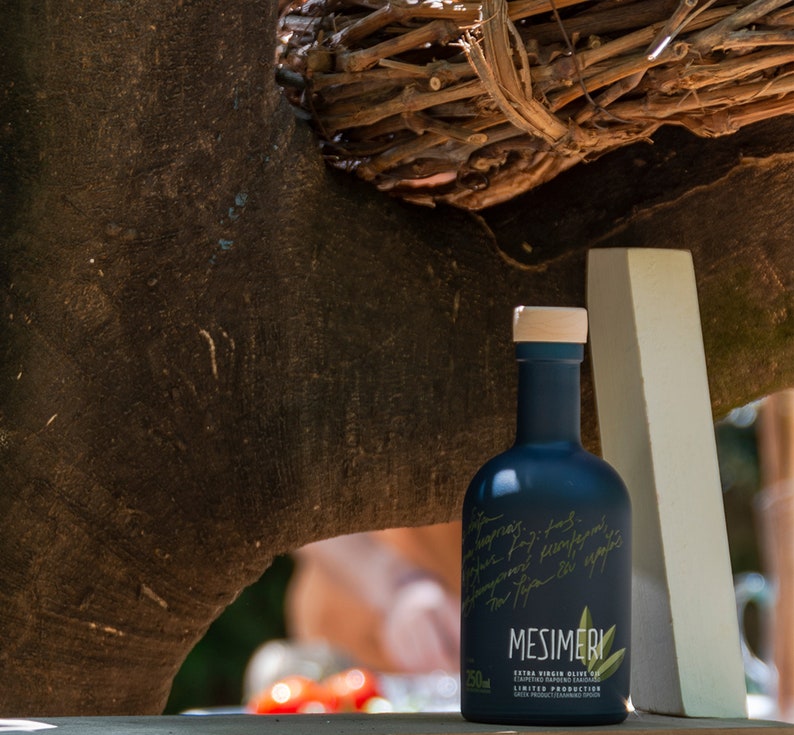 May include: A dark blue glass bottle of Mesimeri Extra Virgin Olive Oil with a white label and handwritten text. The bottle is sitting on a white surface with a wooden background.