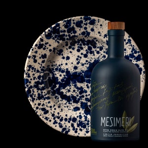 Op de afbeelding: Een donkerblauwe glazen fles Mesimeri Extra Vierge Olijfolie met een houten dop. De fles is gelabeld met de merknaam en de woorden "Extra Vierge Olijfolie" en "Beperkte Productie". De fles staat op een wit keramiek bord met blauwe spatten.