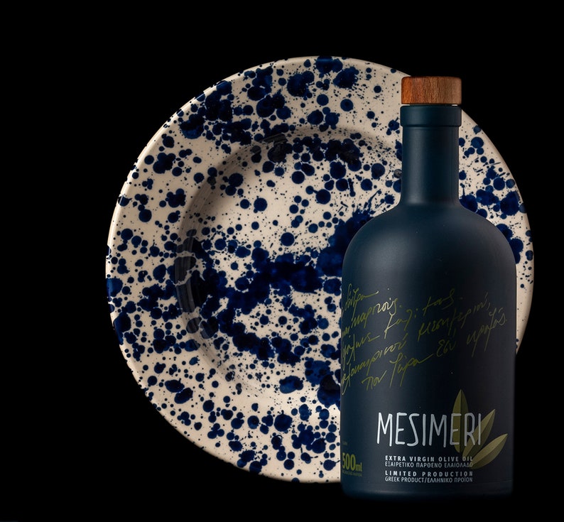 Op de afbeelding: Een donkerblauwe glazen fles Mesimeri Extra Vierge Olijfolie met een houten dop. De fles is gelabeld met de merknaam en de woorden "Extra Vierge Olijfolie" en "Beperkte Productie". De fles staat op een wit keramiek bord met blauwe spatten.