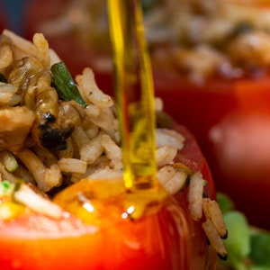 May include: Close-up of a red tomato stuffed with rice, vegetables, and herbs. A clear, yellow liquid is being poured over the tomato.