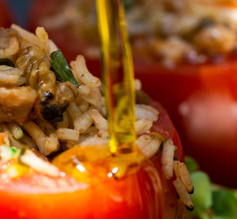 May include: Close-up of a red tomato stuffed with rice, vegetables, and herbs. A clear, yellow liquid is being poured over the tomato.