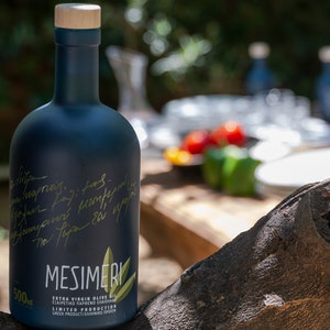 Op de afbeelding: Een donkerblauwe glazen fles Mesimeri Extra Vierge Olijfolie met een houten dop. De fles is gelabeld met de merknaam en de woorden "Extra Vierge Olijfolie" en "Beperkte Productie". De fles staat op een houten oppervlak.
