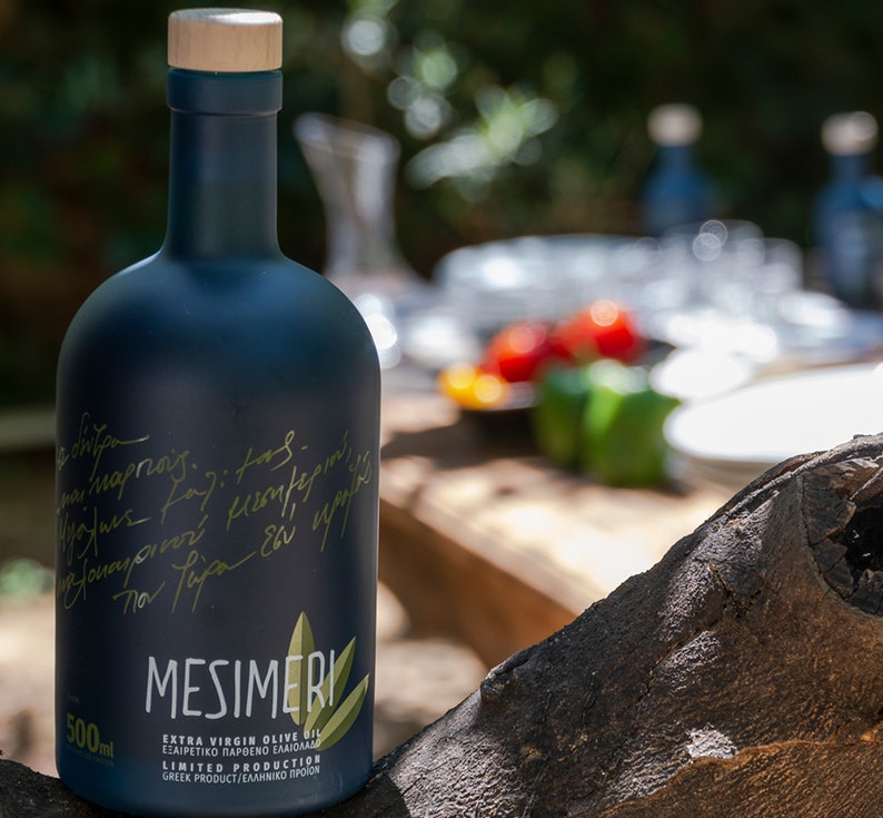 Op de afbeelding: Een donkerblauwe glazen fles Mesimeri Extra Vierge Olijfolie met een houten dop. De fles is gelabeld met de merknaam en de woorden "Extra Vierge Olijfolie" en "Beperkte Productie". De fles staat op een houten oppervlak.