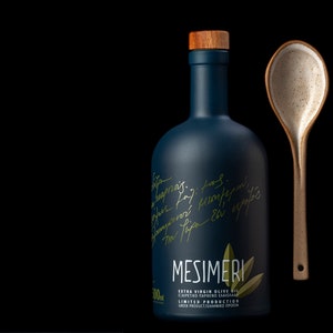 Op de afbeelding: Een donkerblauwe glazen fles met een houten dop en een gouden etiket met de tekst "MESIMERI EXTRA VIRGIN OLIVE OIL &Epsilon;&Xi;&Alpha;&Iota;&Rho;&Epsilon;&Tau;&Iota;&Kappa;&Omicron; &Pi;&Alpha;&Rho;&Theta;&Epsilon;&Nu;&Omicron; &Epsilon;&Lambda;&Alpha;&Iota;&Omicron;&Lambda;&Alpha;&Delta;&Omicron; LIMITED PRODUCTION GREEK PRODUCT/&Epsilon;&Lambda;&Lambda;&Eta;&Nu;&Iota;&Kappa;&Omicron; &Pi;&Rho;&Omicron;Ϊ&Omicron;&Nu;". De fles staat naast een gespikkelde keramische lepel.