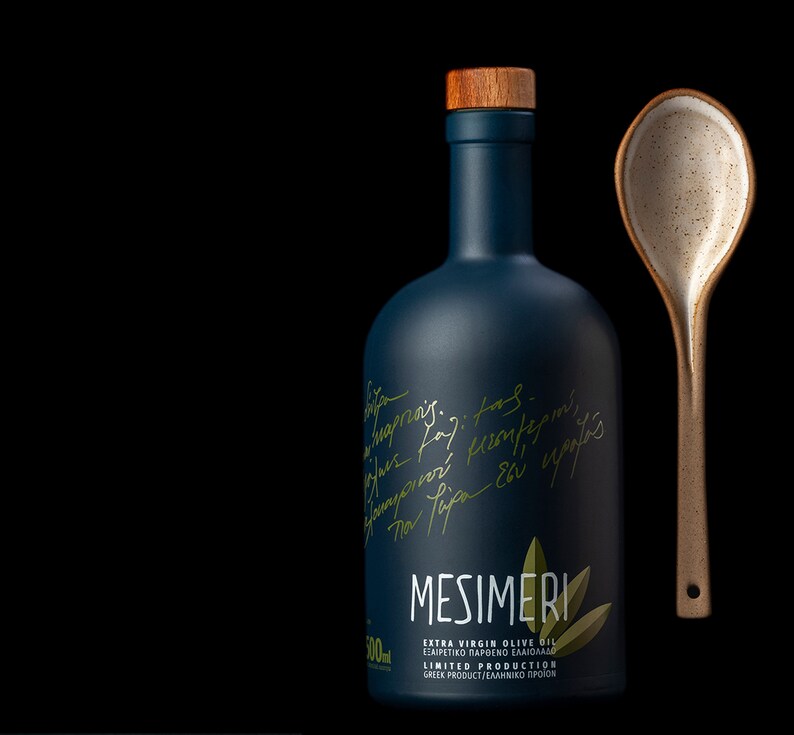 Op de afbeelding: Een donkerblauwe glazen fles met een houten dop en een gouden etiket met de tekst "MESIMERI EXTRA VIRGIN OLIVE OIL &Epsilon;&Xi;&Alpha;&Iota;&Rho;&Epsilon;&Tau;&Iota;&Kappa;&Omicron; &Pi;&Alpha;&Rho;&Theta;&Epsilon;&Nu;&Omicron; &Epsilon;&Lambda;&Alpha;&Iota;&Omicron;&Lambda;&Alpha;&Delta;&Omicron; LIMITED PRODUCTION GREEK PRODUCT/&Epsilon;&Lambda;&Lambda;&Eta;&Nu;&Iota;&Kappa;&Omicron; &Pi;&Rho;&Omicron;Ϊ&Omicron;&Nu;". De fles staat naast een gespikkelde keramische lepel.