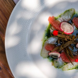 May include: A white plate with a seafood dish featuring thin slices of raw fish, grapefruit segments, radish slices, and green seaweed. The dish is drizzled with a green sauce and garnished with red flower petals.