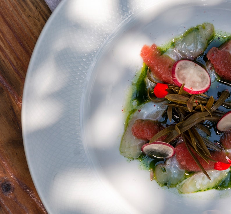 May include: A white plate with a seafood dish featuring thin slices of raw fish, grapefruit segments, radish slices, and green seaweed. The dish is drizzled with a green sauce and garnished with red flower petals.