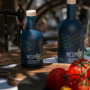 May include: Two dark blue glass bottles of Mesimeri Extra Virgin Olive Oil, one 500 ml bottle and one smaller bottle, on a wooden cutting board with tomatoes and green peppers.