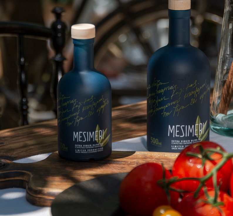 May include: Two dark blue glass bottles of Mesimeri Extra Virgin Olive Oil, one 500 ml bottle and one smaller bottle, on a wooden cutting board with tomatoes and green peppers.