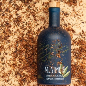 Op de afbeelding: Een donkerblauwe glazen fles Mesime extra vierge olijfolie met een houten dop. De fles is gelabeld met de merknaam "Mesime" en de tekst "Extra vierge olijfolie", "Beperkte productie" en "Grieks product/&Epsilon;&lambda;&lambda;&eta;&nu;&iota;&kappa;ό &Pi;&rho;&omicron;ϊό&nu;".