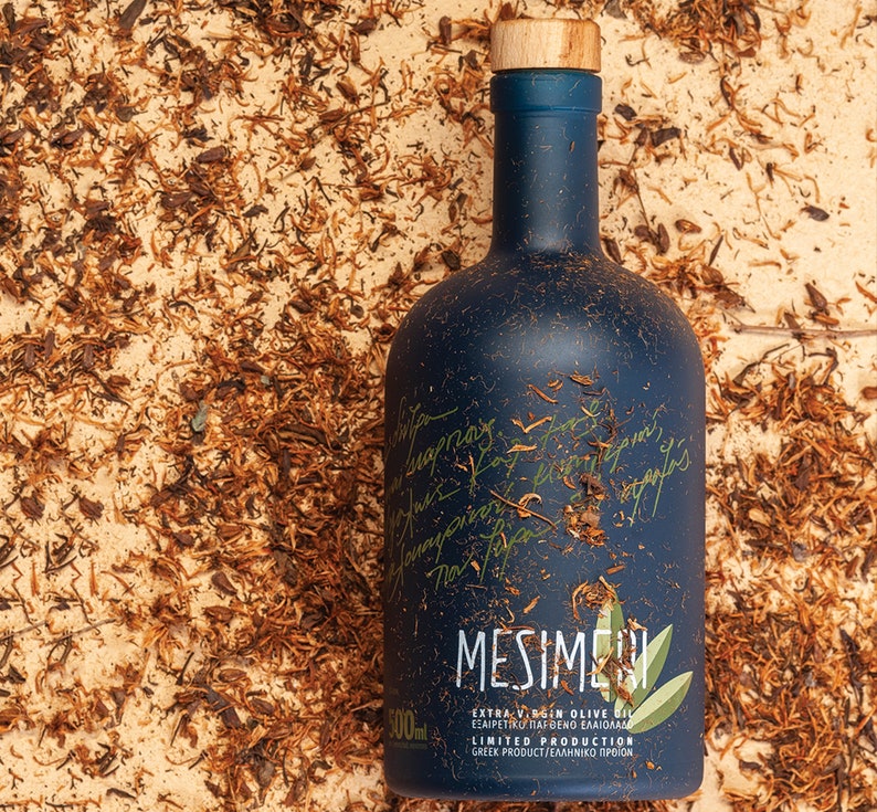 Op de afbeelding: Een donkerblauwe glazen fles Mesime extra vierge olijfolie met een houten dop. De fles is gelabeld met de merknaam "Mesime" en de tekst "Extra vierge olijfolie", "Beperkte productie" en "Grieks product/&Epsilon;&lambda;&lambda;&eta;&nu;&iota;&kappa;ό &Pi;&rho;&omicron;ϊό&nu;".