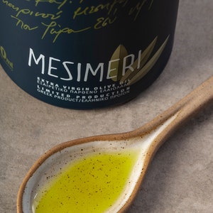 May include: A dark blue bottle of Mesimeri Extra Virgin Olive Oil with a gold leaf design. The bottle is labeled with the text "Mesimeri Extra Virgin Olive Oil", "&Epsilon;&xi;&alpha;&iota;&rho;&epsilon;&tau;&iota;&kappa;ό &Pi;&alpha;&rho;&theta;έ&nu;&omicron; &Epsilon;&lambda;&alpha;&iota;ό&lambda;&alpha;&delta;&omicron;", "Limited Production", and "Greek Product/&Epsilon;&lambda;&lambda;&eta;&nu;&iota;&kappa;ό &Pi;&rho;&omicron;ϊό&nu;".