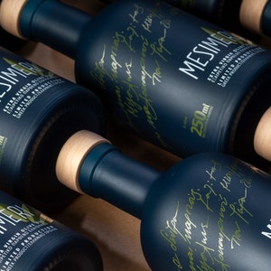 May include: Close-up of four dark blue glass bottles with wooden caps. Each bottle is labeled with the text "Mesimeri Extra Virgin Olive Oil" in Greek and English. The bottles are arranged in a row on a wooden surface.