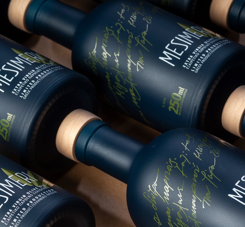 May include: Close-up of four dark blue glass bottles with wooden caps. Each bottle is labeled with the text "Mesimeri Extra Virgin Olive Oil" in Greek and English. The bottles are arranged in a row on a wooden surface.
