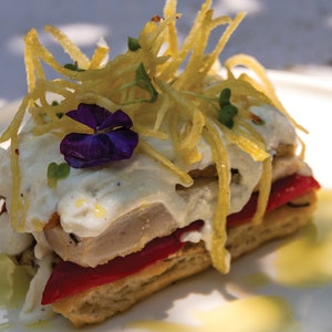 Op de afbeelding: Een gelaagd gerecht met een witte saus, rode paprika's en een paarse bloem erop. Het gerecht is gegarneerd met knapperige aardappelstroken.