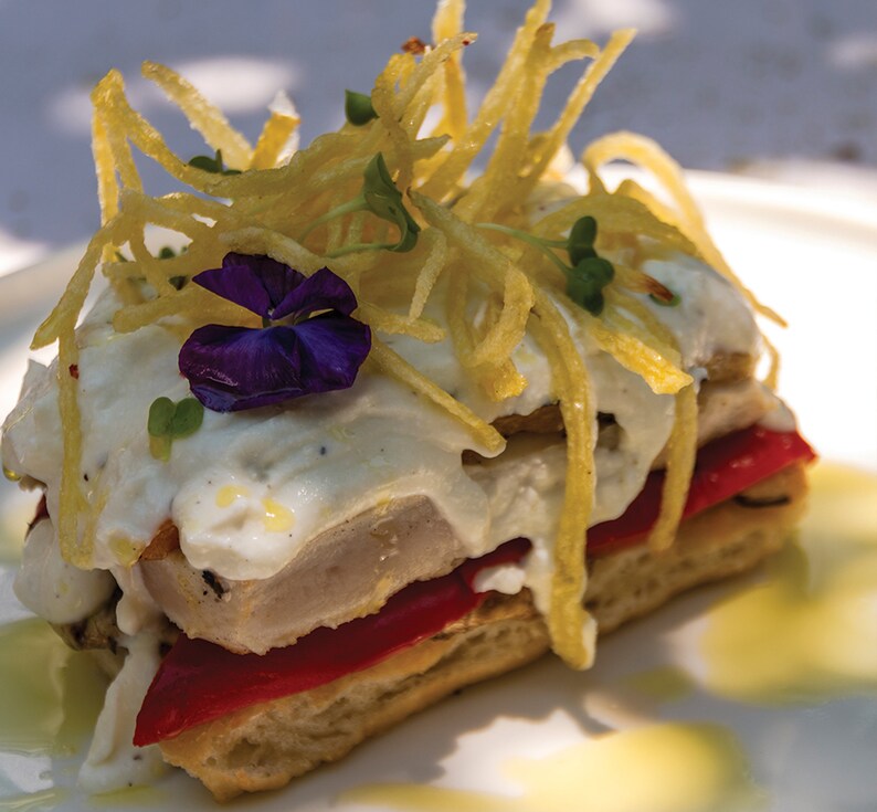 Op de afbeelding: Een gelaagd gerecht met een witte saus, rode paprika's en een paarse bloem erop. Het gerecht is gegarneerd met knapperige aardappelstroken.
