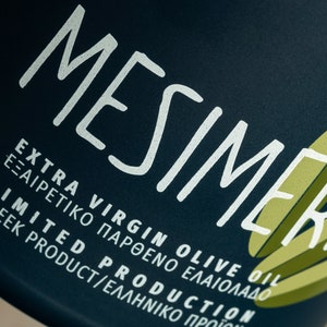 Op de afbeelding: Een donkerblauwe fles met witte tekst die "MESIMER EXTRA VIRGIN OLIVE OIL &Epsilon;&Xi;&Alpha;&Iota;&Rho;&Epsilon;&Tau;&Iota;&Kappa;&Omicron; &Pi;&Alpha;&Rho;&Theta;&Epsilon;&Nu;&Omicron; &Epsilon;&Lambda;&Alpha;&Iota;&Omicron;&Lambda;&Alpha;&Delta;&Omicron; LIMITED PRODUCTION GREEK PRODUCT/&Epsilon;&Lambda;&Lambda;&Eta;&Nu;&Iota;&Kappa;&Omicron; &Pi;&Rho;&Omicron;Ϊ&Omicron;&Nu;" zegt.