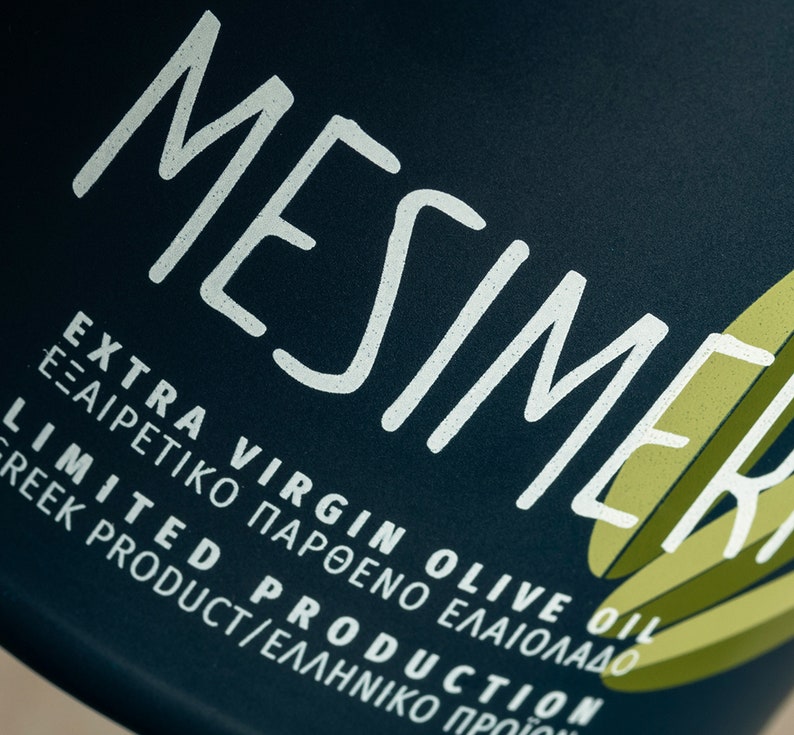 Op de afbeelding: Een donkerblauwe fles met witte tekst die "MESIMER EXTRA VIRGIN OLIVE OIL &Epsilon;&Xi;&Alpha;&Iota;&Rho;&Epsilon;&Tau;&Iota;&Kappa;&Omicron; &Pi;&Alpha;&Rho;&Theta;&Epsilon;&Nu;&Omicron; &Epsilon;&Lambda;&Alpha;&Iota;&Omicron;&Lambda;&Alpha;&Delta;&Omicron; LIMITED PRODUCTION GREEK PRODUCT/&Epsilon;&Lambda;&Lambda;&Eta;&Nu;&Iota;&Kappa;&Omicron; &Pi;&Rho;&Omicron;Ϊ&Omicron;&Nu;" zegt.