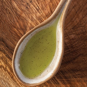 May include: A small, white ceramic spoon filled with a light green liquid. The spoon is sitting on a wooden surface.