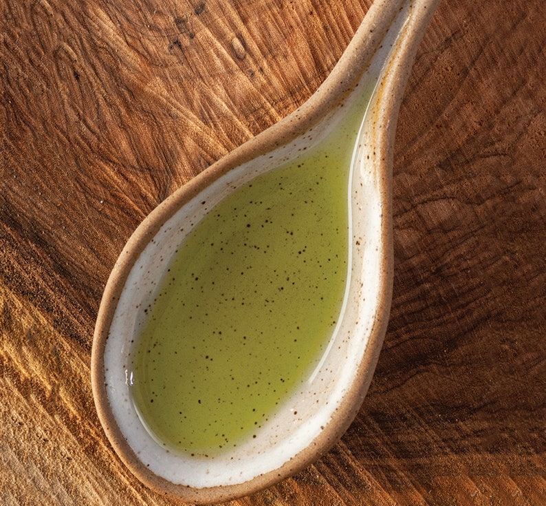 May include: A small, white ceramic spoon filled with a light green liquid. The spoon is sitting on a wooden surface.