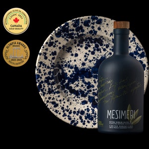 May include: A dark blue glass bottle of Mesimeri Extra Virgin Olive Oil with a gold label and a wooden stopper. The bottle is sitting on a white and blue ceramic plate with a speckled pattern.