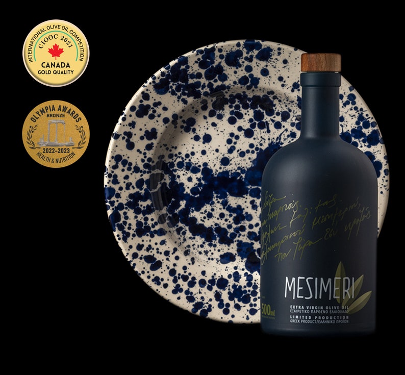 May include: A dark blue glass bottle of Mesimeri Extra Virgin Olive Oil with a gold label and a wooden stopper. The bottle is sitting on a white and blue ceramic plate with a speckled pattern.