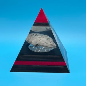 Peut inclure: Un presse-papier en résine transparent en forme de pyramide, avec un sommet rouge et une base noire. Un modèle blanc d'un vaisseau spatial est suspendu à l'intérieur. Fond bleu vif.