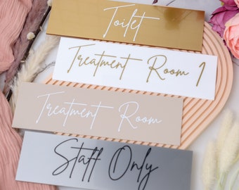 Puede incluir: Cuatro letreros rectangulares con diferentes colores y texto. El letrero superior es dorado con texto blanco que dice "Toilette". El segundo letrero es blanco con texto dorado que dice "Sala de tratamiento 1". El tercer letrero es beige con texto blanco que dice "Sala de tratamiento". El letrero inferior es gris con texto negro que dice "Solo personal".