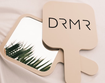 May include: Two beige hand mirrors with square-shaped reflective surfaces and handles. The word "DRMR" is printed in black on one mirror. A gold candle holder is in the background.