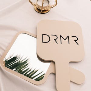 Könnte beinhalten: Zwei beige Handspiegel mit quadratischen Spiegelflächen und Griffen. Das Wort "DRMR" ist schwarz auf einem Spiegel aufgedruckt. Ein goldener Kerzenhalter befindet sich im Hintergrund.