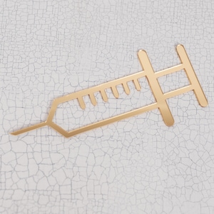 Puede incluir: Un objeto decorativo en forma de jeringa de color dorado. La jeringa tiene un diseño detallado con una aguja y un émbolo. El objeto está colocado sobre una superficie blanca y agrietada, creando un contraste.