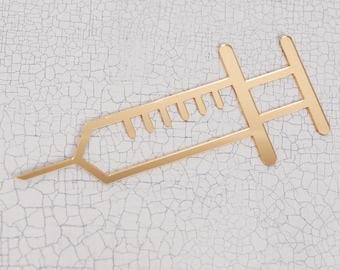 May include: A gold-colored syringe-shaped decorative item. The syringe has a detailed design with a needle and a plunger. The item is placed on a white, cracked surface, creating a contrast between the smooth metal and the textured background.