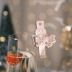 May include: Rose gold nutcracker shaped drink charm with the text "ink etch" on the side. The charm is hanging from a glass of champagne with a strawberry inside.