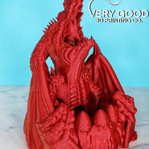 May include: A 3D printed red dragon figurine with its mouth open and wings spread. The dragon is sitting on a nest of eggs.