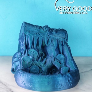Puede incluir: Un cofre del tesoro impreso en 3D de color azul turquesa con la boca de un monstruo abierta, revelando monedas de oro en su interior. El cofre está sentado sobre una superficie blanca.  "VERY GOOD 3D PRINTING CO." está impreso en el fondo.