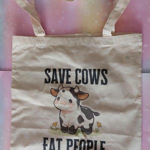 K&ouml;nnte beinhalten: Beige Canvas-Tragetasche mit dem schwarzen Text "SAVE COWS EAT PEOPLE". Die Tasche zeigt eine Cartoon-Kuh-Illustration mit schwarzen Flecken und einer rosa Nase. Die Tasche hat zwei beige Griffe.