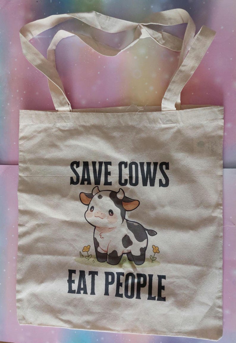 K&ouml;nnte beinhalten: Beige Canvas-Tragetasche mit dem schwarzen Text "SAVE COWS EAT PEOPLE". Die Tasche zeigt eine Cartoon-Kuh-Illustration mit schwarzen Flecken und einer rosa Nase. Die Tasche hat zwei beige Griffe.