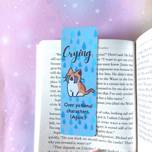 Könnte beinhalten: Ein blaues und weißes Lesezeichen mit einer Comic-Katze, die eine Brille trägt und weint. Das Lesezeichen hat den Text "Crying. Over fictional characters. (Again.)" und ein Muster aus Regentropfen.