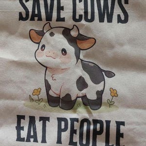 K&ouml;nnte beinhalten: Eine Tragetasche aus Segeltuch mit dem Text "SAVE COWS EAT PEOPLE" in Schwarz. Die Tasche zeigt eine Cartoon-Illustration einer schwarz-wei&szlig;en Kuh mit rosa Rouge und kleinen gelben Blumen.